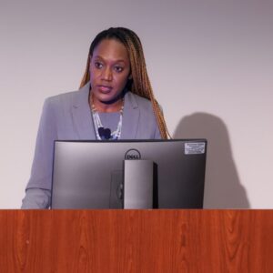 Dr. Rollins, a woman with long braided hair wearing a gray blazer is standing behind a wooden podium, speaking at the 2024 NAACFRC Annual Conference. She is looking attentively at the audience while standing in front of a computer monitor.