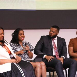 A panel of four people seated on stage at a conference. The woman on the left, wearing a white blazer, listens attentively. The woman next to her, wearing a patterned dress, is speaking into a microphone. The man next to her in a pinstripe suit and patterned tie looks on thoughtfully. The woman on the far right, wearing a brown dress, is also listening intently.